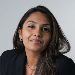 iuGO Digital - Woman with long dark hair wearing a black blazer and gold hoop earrings, looking at the camera with a neutral expression against a plain background.