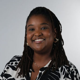 iuGO Digital - A woman with long, dark braided hair and gold hoop earrings smiles at the camera. She is wearing a black and white patterned shirt and a necklace. The background is plain light gray.