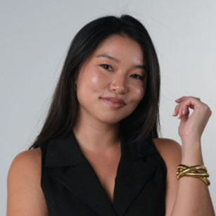 iuGO Digital - A woman with long dark hair, wearing a sleeveless black top and gold bracelet, stands against a plain light background, smiling gently at the camera.