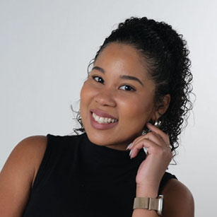iuGO Digital - Woman with curly hair wearing a sleeveless black top and a watch, smiling and touching her face, posed against a plain light background.