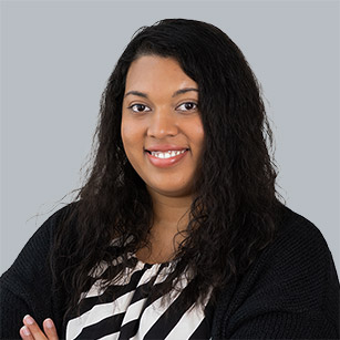 iuGO Digital - Woman with long curly hair and a black sweater over a black-and-white striped blouse, smiling with arms crossed, posed in front of a plain gray background.