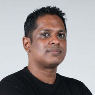 iuGO Digital - A man with short black hair wearing a black t-shirt stands against a plain light gray background, looking at the camera with a neutral expression.