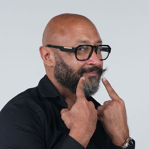 iuGO Digital - A bald man with a beard and glasses points to his mustache with both index fingers, wearing a black shirt against a plain light background.
