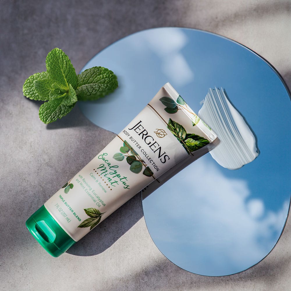 iuGO Digital - Jergens Eucalyptus Mint body butter tube on a reflective surface with a mint sprig and a smear of white cream.